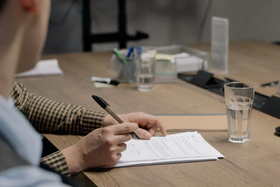 Personne en tenue professionnelle signant un document sur une table en bois dans un cadre de bureau.