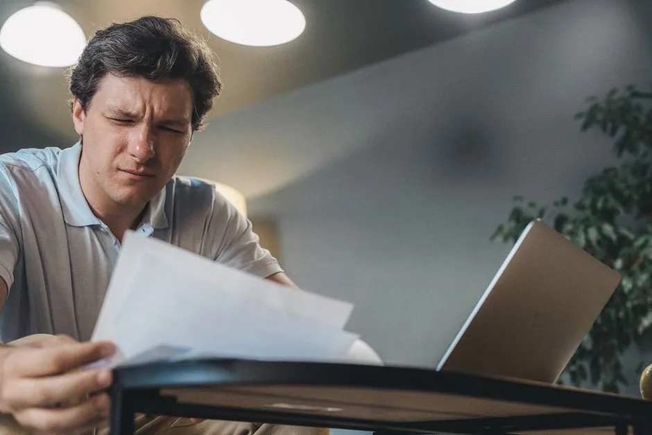 Homme se concentrant intensément tout en examinant des documents à un bureau avec un ordinateur portable.